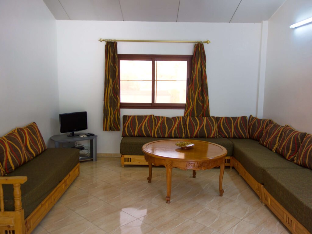 A simple living room with patterned cushions on two wooden sofas, a round wooden coffee table in the center, tiled floor, and matching curtains—perfect for relaxing after a day exploring Camping-Aourir Taghazout Agdir.