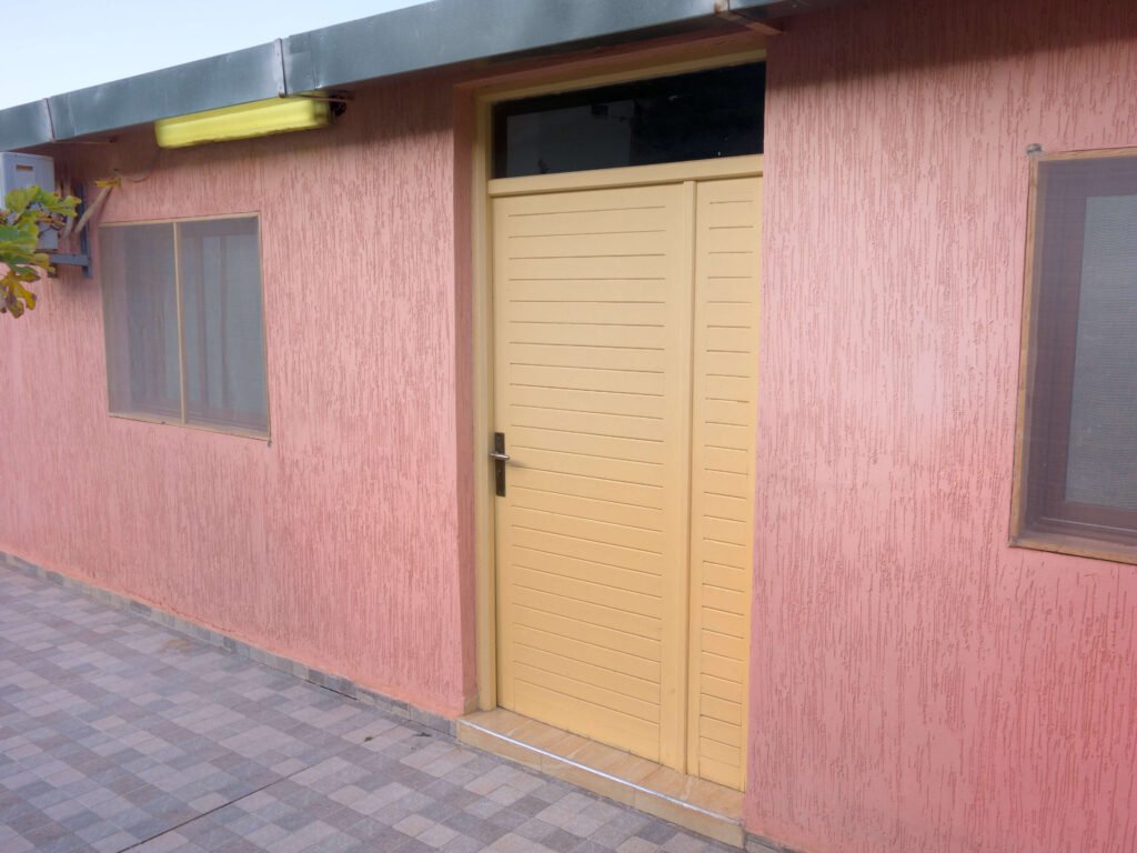A closed yellow door set in a pink textured wall at Camping-Aourir Taghazout Agdir, with a small rectangular window above the door and another window to the left. The floor is tiled with a gray and beige checkered pattern.