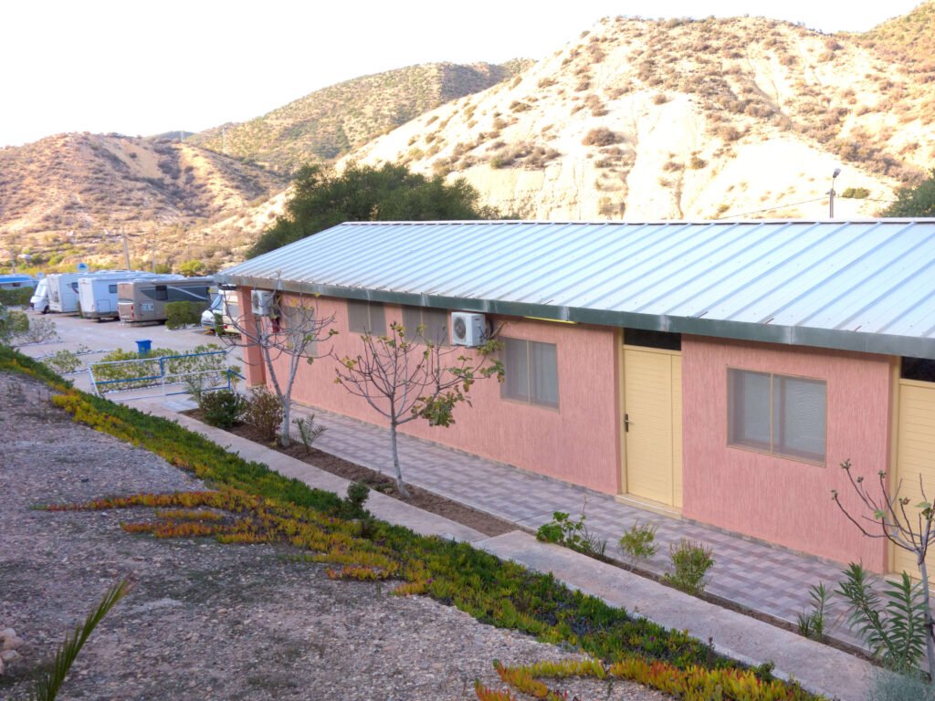 A long, single-story pink building with a metal roof and yellow doors stands in the dry, hilly landscape of Camping-Aourir Taghazout Agdir. Small trees and low plants line the walkway; trailers are visible in the background.