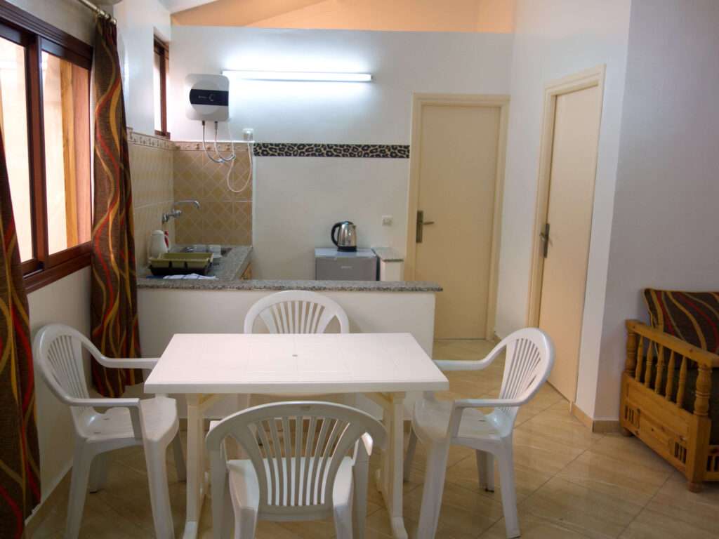 A small kitchen and dining area at Camping-Aourir Taghazout Agdir features a white plastic table with four chairs, a sink and counter, kettle, tiled backsplash, two closed doors, brown-curtained windows, and a wooden bench on the right.