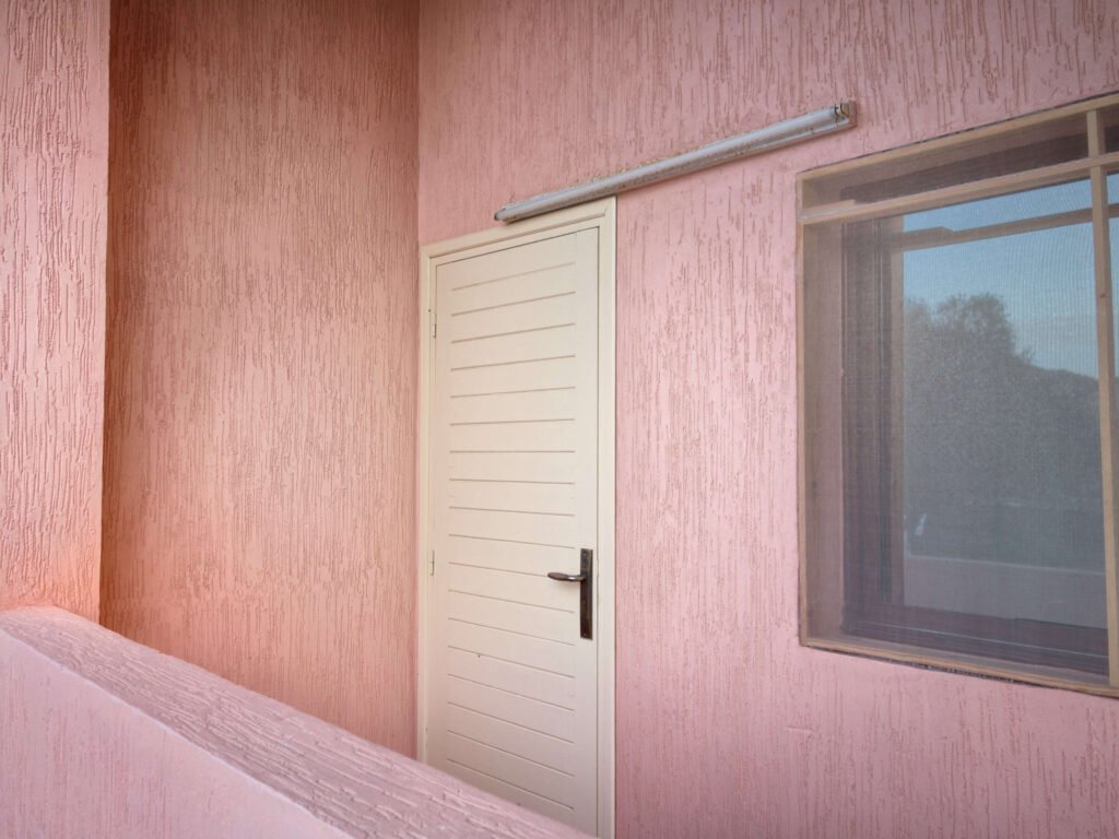 A white door with a black handle is set in a pink textured wall next to a window with a screen; the pink balcony railing overlooks Camping-Aourir Taghazout Agadir.