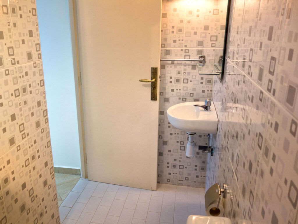 A small bathroom at Camping-Aourir Taghazout Agadir features patterned tile walls, a white sink with faucet, a wall-mounted mirror, towel bar, toilet paper holder, and a partially open white door.