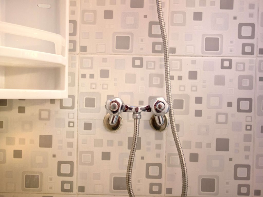 A close-up of a shower wall with a retro square-patterned tile design at Camping-Aourir Taghazout Agadir. A chrome shower faucet with two handles and a flexible hose is mounted below a white plastic shelf.