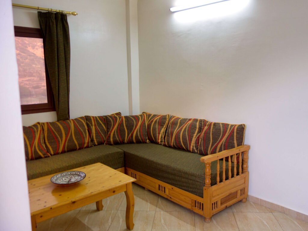 A small living room corner at Camping-Aourir Taghazout Agadir features a wooden sectional sofa with patterned cushions, a matching coffee table topped with a decorative plate, cream-colored tiled floor, and a window dressed in a dark curtain.