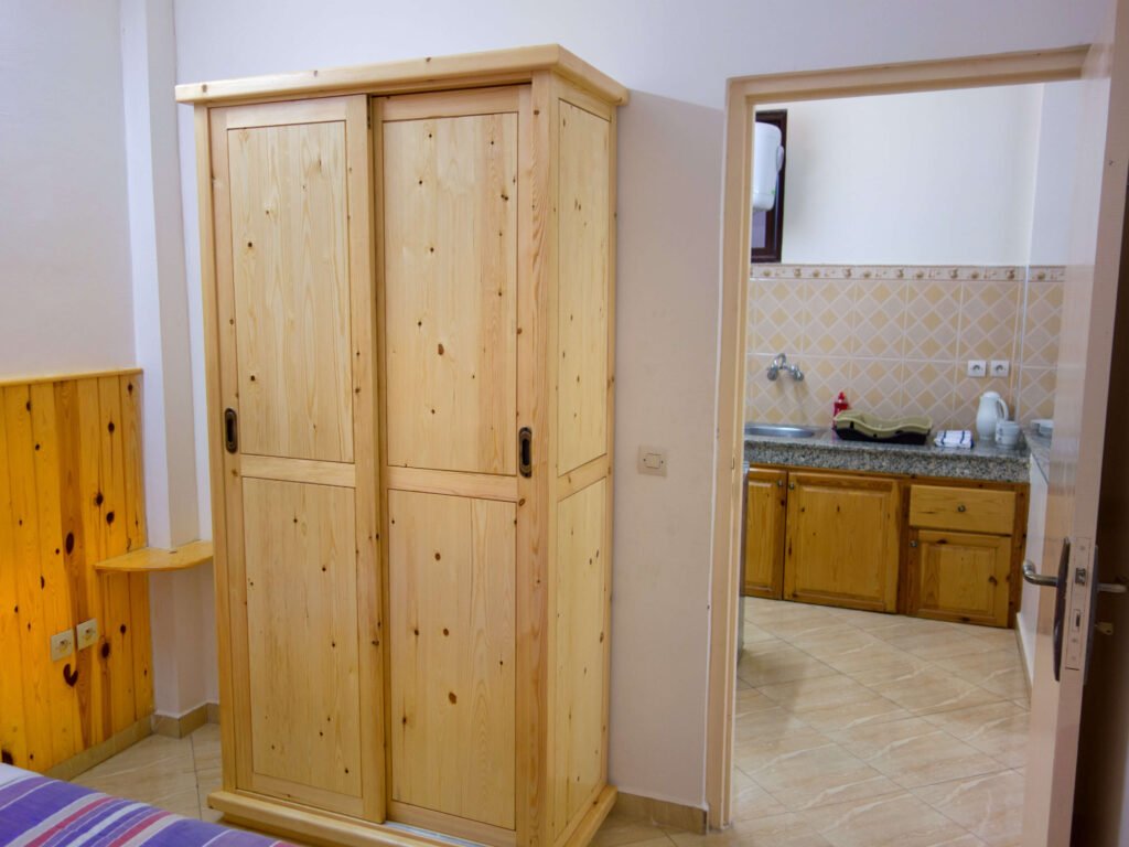 A wooden wardrobe stands in a bedroom next to an open door leading to a kitchen with wooden cabinets, beige tiled floors, and a gray countertop—evoking the cozy style of Camping-Aourir Taghazout Agadir.